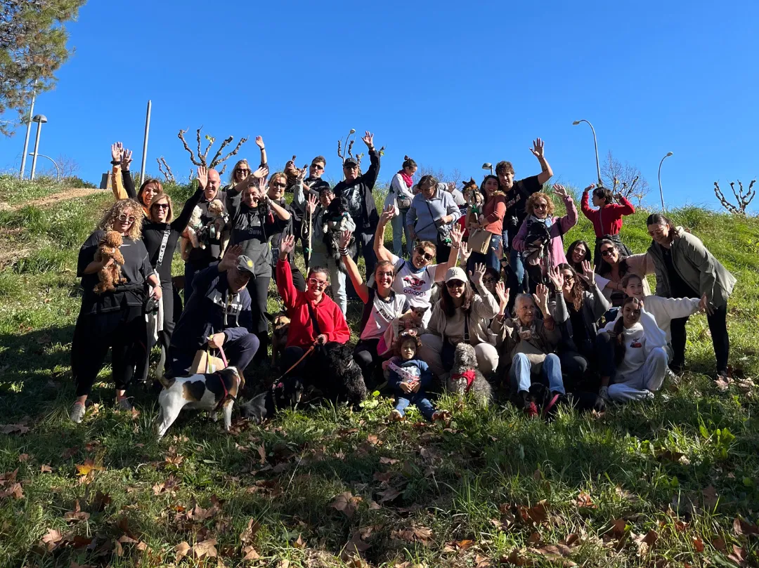 Grup de participants del Gos&Glop a Sant Quirze somrient i saludant durant la caminada, acompanyats dels seus gossos en un matí assolellat al bosc.